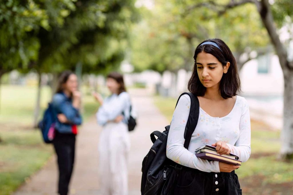 Teenage girl carrying books looking sad with two girls in the bakground in a conversation.