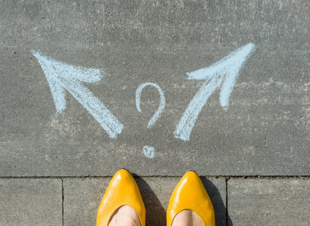 Yellow shoes standing on pavement, in the middle of two chalk drawings of arrows pointing into two different directions, with a question mark drawn in the middle.