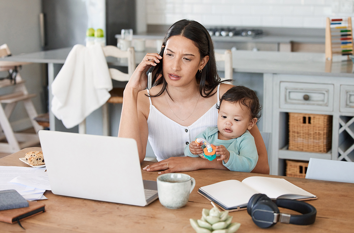 Shot of a woman talking on her cellphone while sitting with her laptop and her baby on her lap.